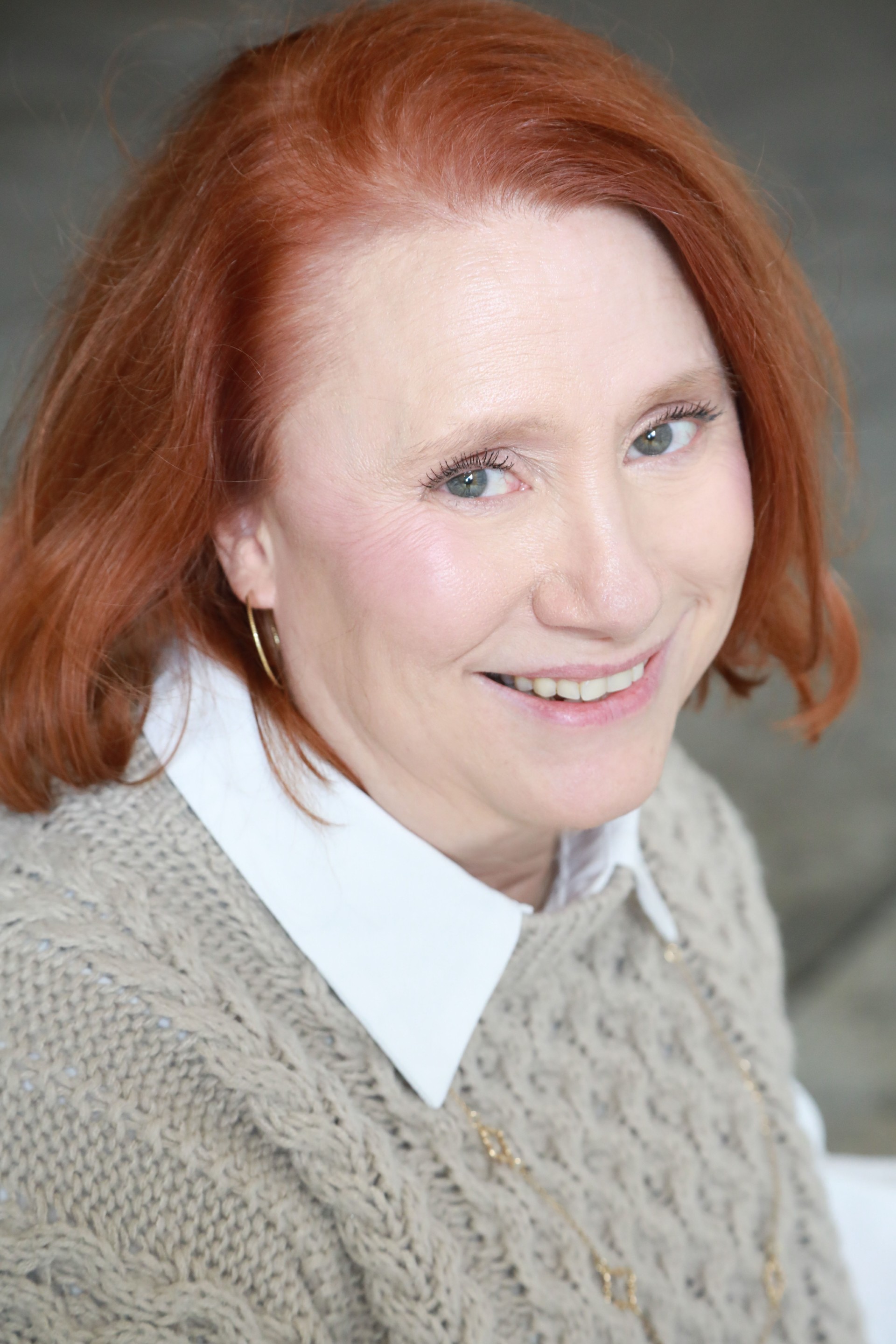 Kimberly Hill Ridley smiling. She has red hair and is wearing a beige sweater over white collared shirt, a gold chain and hooped earrings.