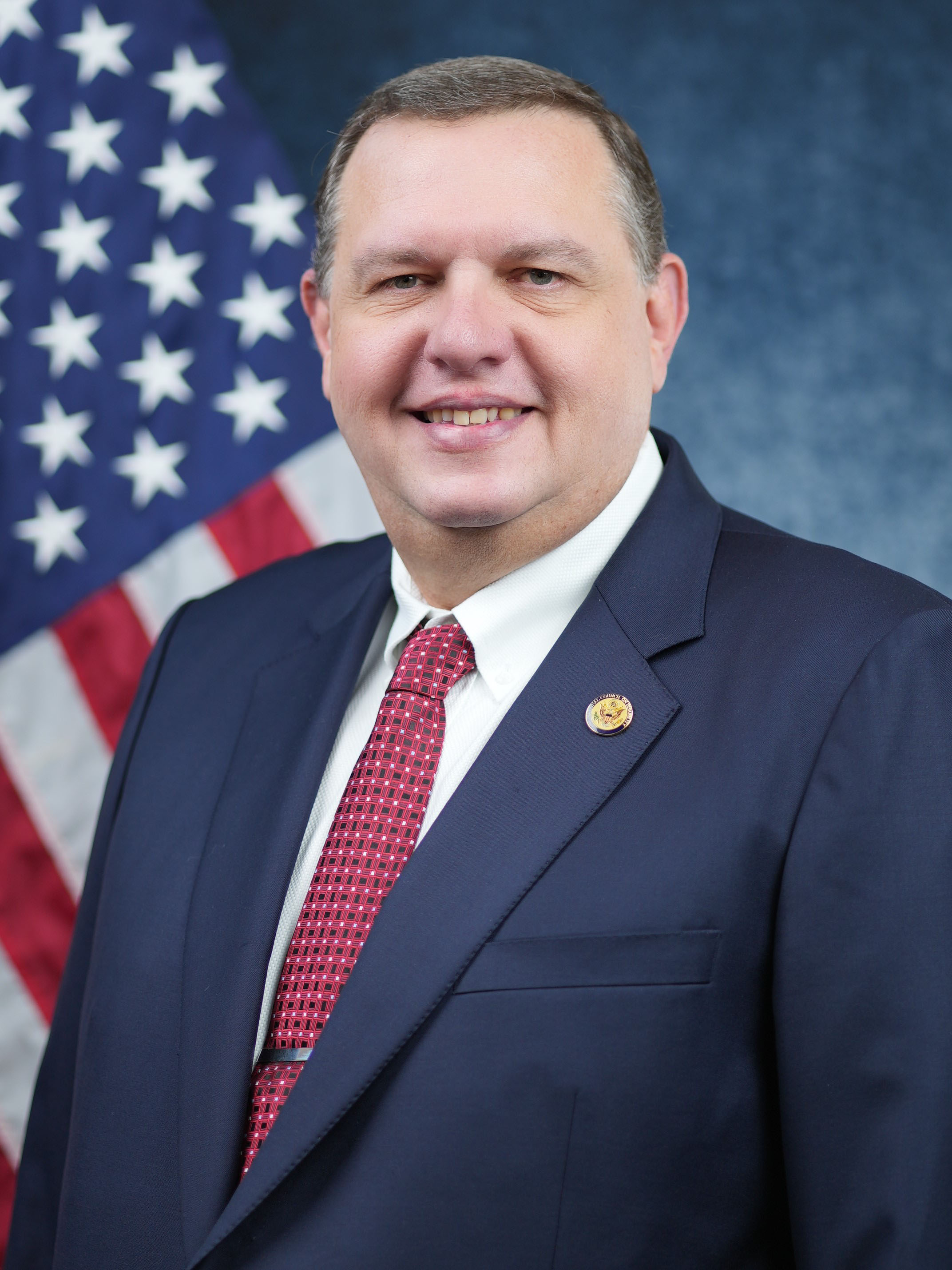 photo of Shawn Kennemer wearing blue suit and red tie with U.S. flag background.